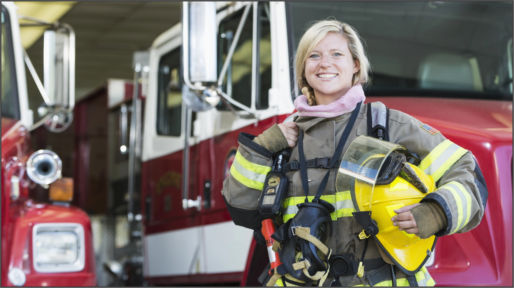 A firefighter in their bunker gear smiles in front of two firetrucks while holding their helmet